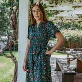 Woman in a floral dress standing outdoors near a white column with greenery in the background