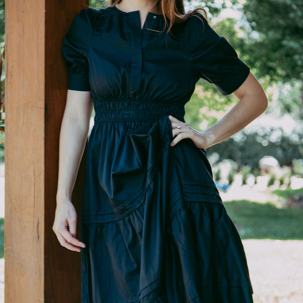 Woman wearing a dark blue dress standing outdoors with greenery in the background