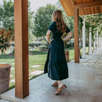 Woman in a green dress standing on a stone patio under a wooden canopy with trees and garden in the background.