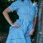 Person wearing a blue and white striped dress leaning against a tree.