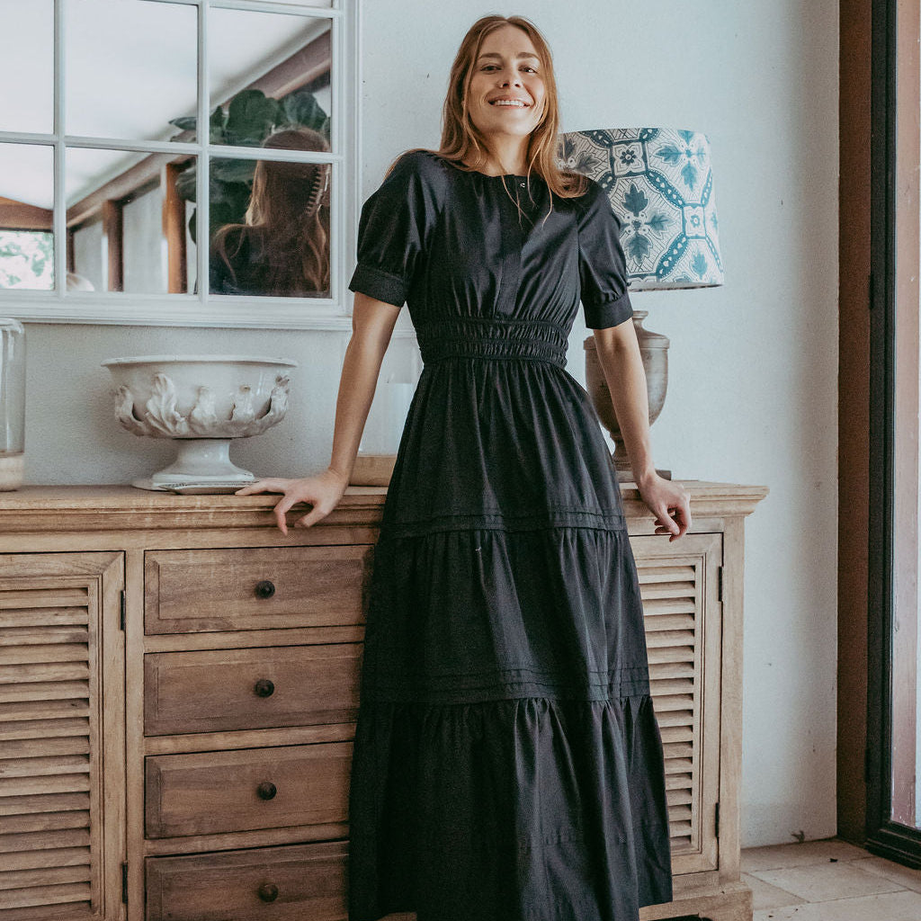 Woman in a black dress standing in a room with wooden furniture and decorative items.