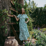 Woman in a floral dress standing on a wooden ladder in a garden.