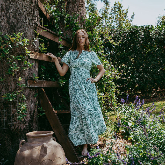 Woman in a floral dress standing on a wooden ladder in a garden.