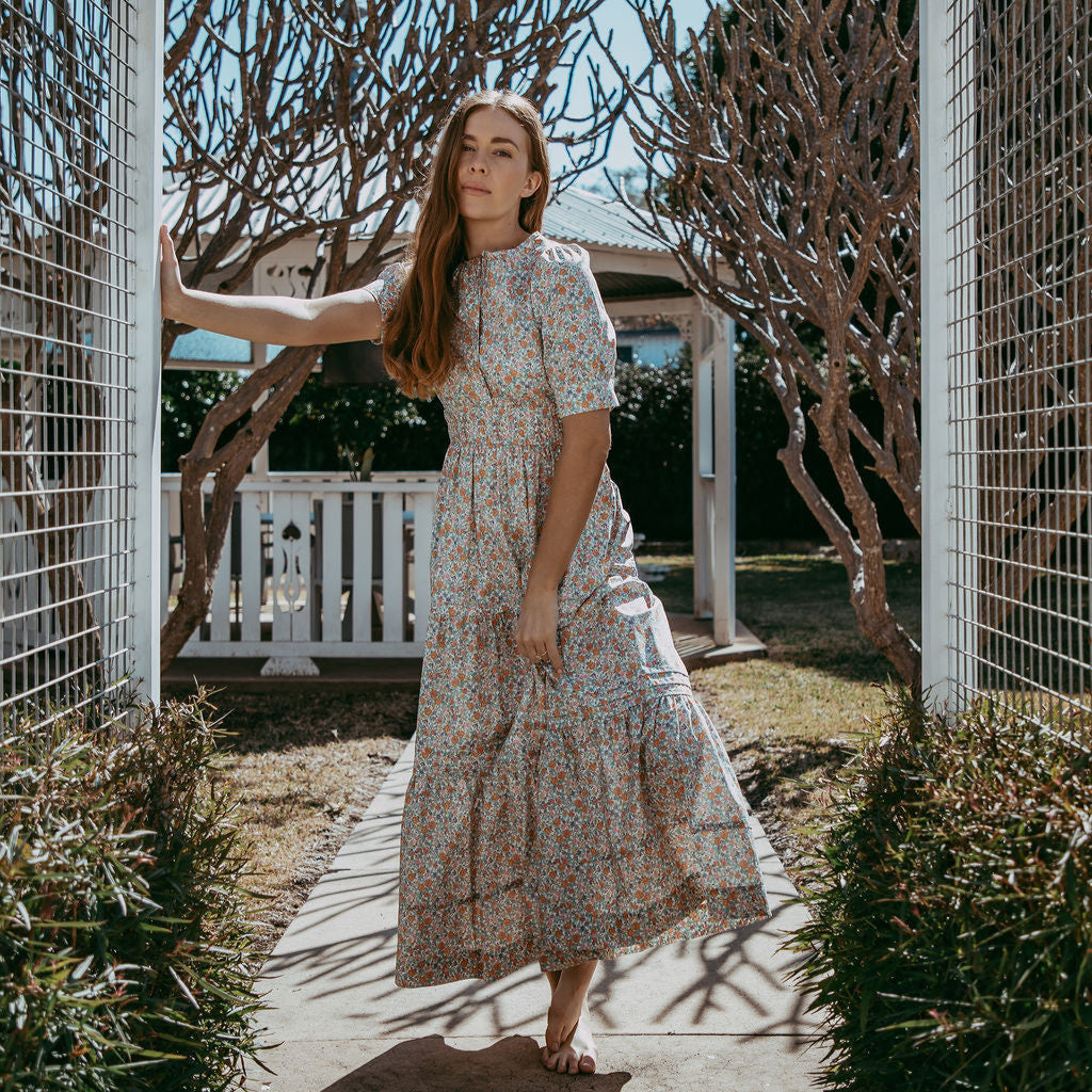 Woman in a floral dress standing in a garden with trees and a fence in the background