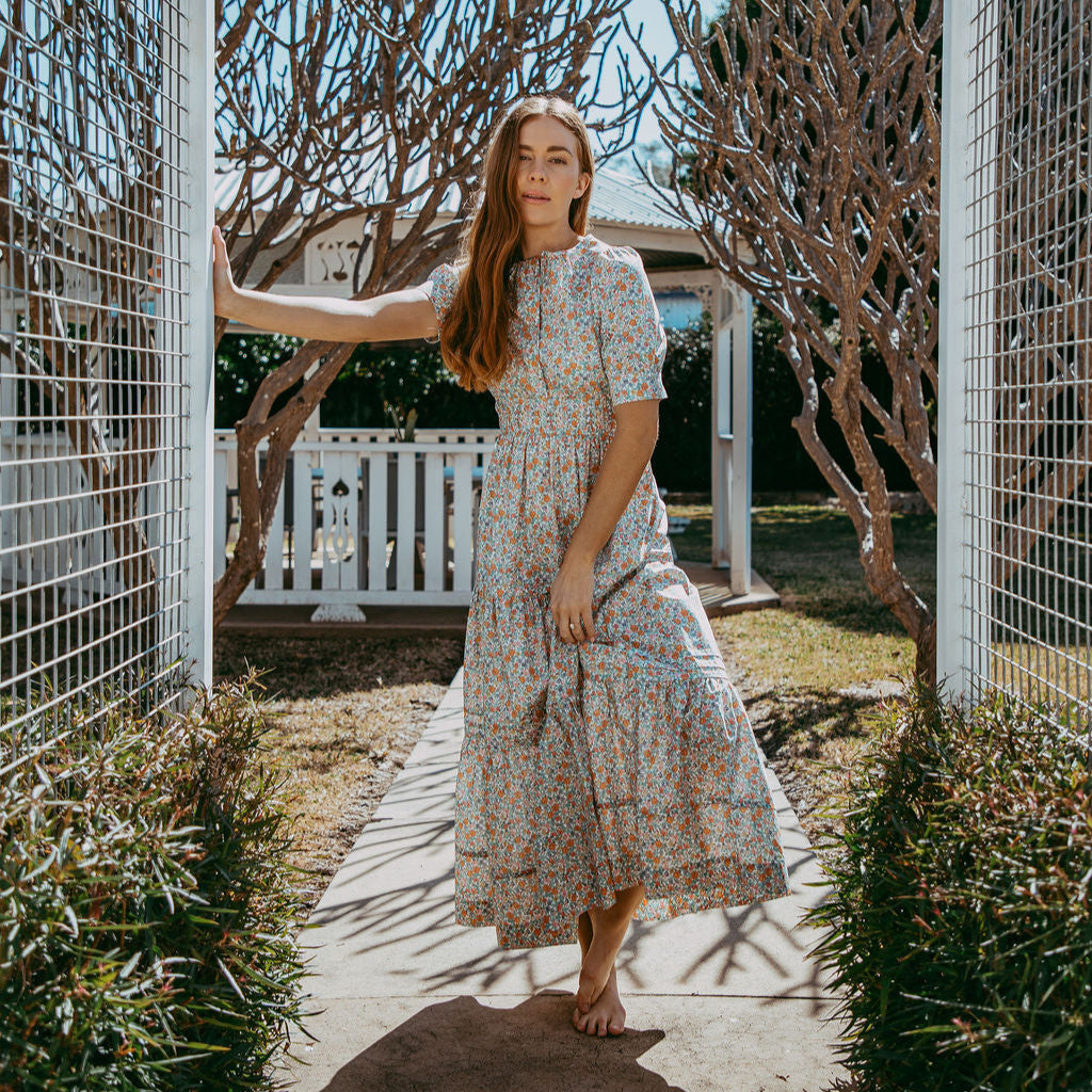 Woman in a floral dress standing in a garden with trees and a white fence in the background.