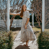 Woman in a floral dress standing in a garden with trees and a white fence in the background.