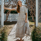 Woman in a floral dress standing in a garden with trees and a white fence in the background.