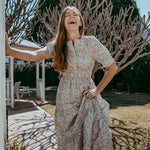 Woman in a floral dress standing outdoors with trees and a house in the background