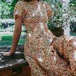 Woman in a floral dress sitting on a wooden bench outdoors with greenery in the background