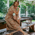 Woman in a floral dress sitting on a wooden bench under a pergola with greenery.