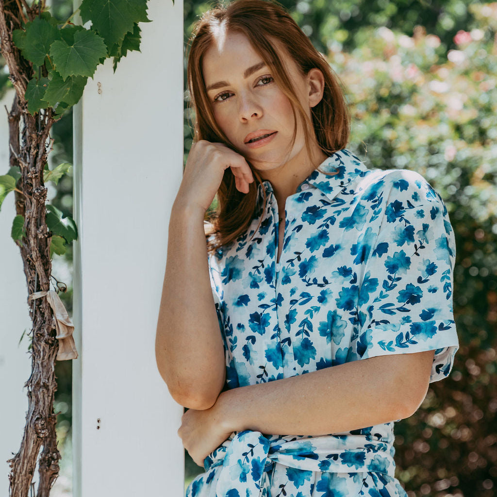 Woman in a blue floral dress leaning against a white column outdoors.