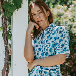 Woman in a blue floral dress leaning against a white column outdoors.