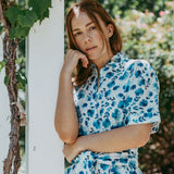 Woman in a blue floral dress leaning against a white column outdoors.
