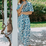 Woman in a floral dress standing under a white pergola with greenery around.