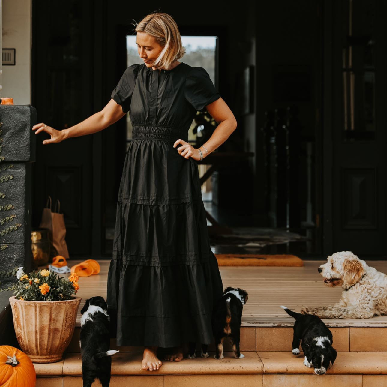 Woman in a black dress standing on a porch with dogs and pumpkins.