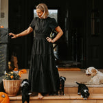Woman in a black dress standing on a porch with dogs and pumpkins.