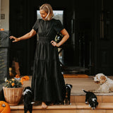 Woman in a black dress standing on a porch with dogs and pumpkins.